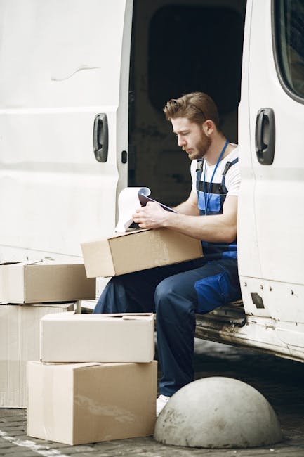 Inside a residential room during a home relocation process, a person is holding a cardboard box labeled 'BOOKS' while standing near a stack of boxes of various sizes, some marked with labels like 'MED