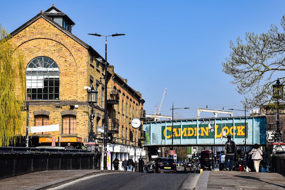 The image shows a train station platform during daylight hours with an industrial-style metal canopy overhead, supported by blue-painted metal beams and trusses. The station building, made of brick wi