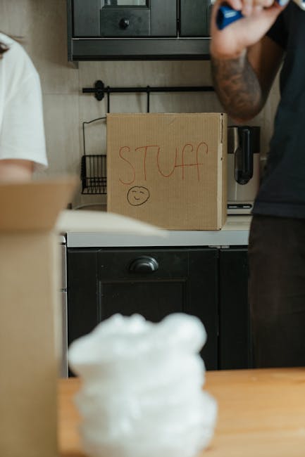 Inside a kitchen area during a home relocation, a cardboard box labeled 'stuf' with a smiley face drawn on it is placed on a white countertop above black kitchen cabinets. A person wearing a dark shir