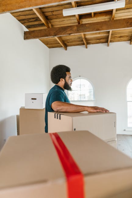 Inside a kitchen area during a home relocation, a cardboard box labeled 'stuf' with a smiley face drawn on it is placed on a white countertop above black kitchen cabinets. A person wearing a dark shir